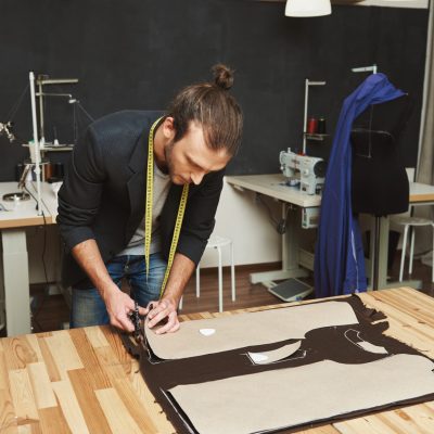 Mature creative handsome dark-haired caucasian male fashion designer in black suit cutting out clothes parts from fabric with scissors, spending evening in studio. Bachelor of Fashion Designing